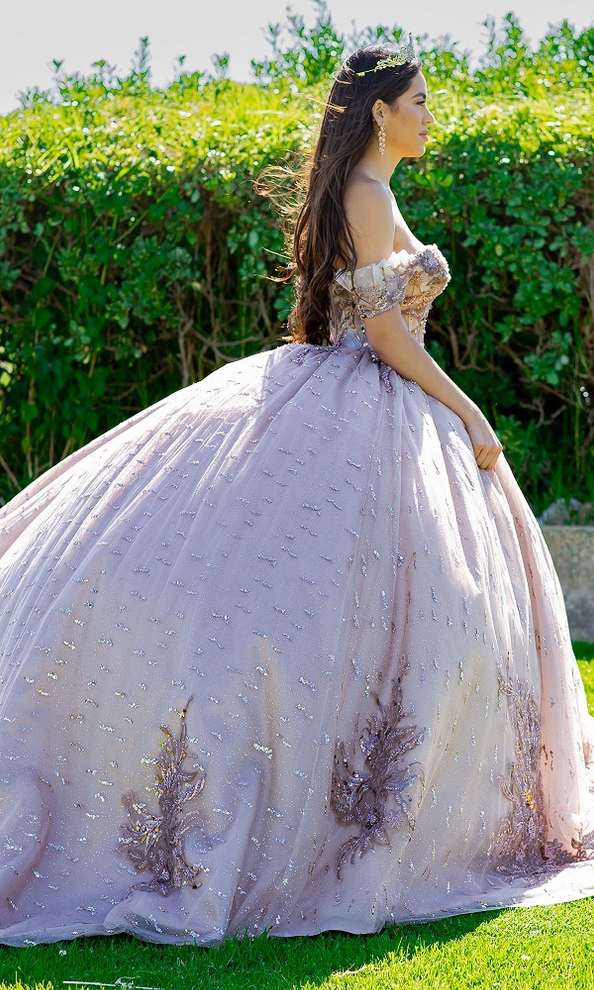 Wearing the Quinceanera Ball Gown 8087J and a tiara, a woman stands on grass with lush greenery behind her.